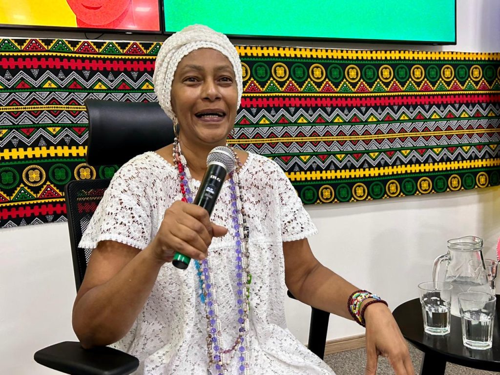 Uma mulher sorrindo, usando turbante branco, vestido de renda e longos colares coloridos, falando ao microfone com um painel de tecido afro-brasileiro ao fundo.
